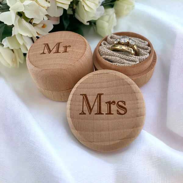 Wooden ring boxes labeled 'Mr' and 'Mrs' with a gold ring inside, on a white fabric background.