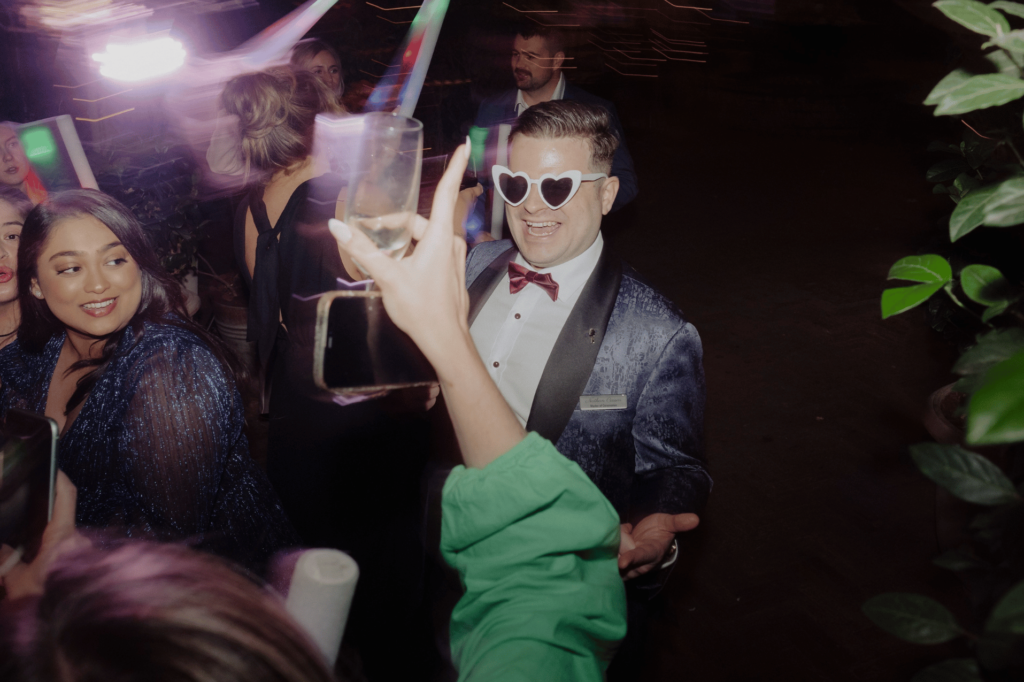 Man in formal attire with bow tie and sunglasses at a party, surrounded by people.
