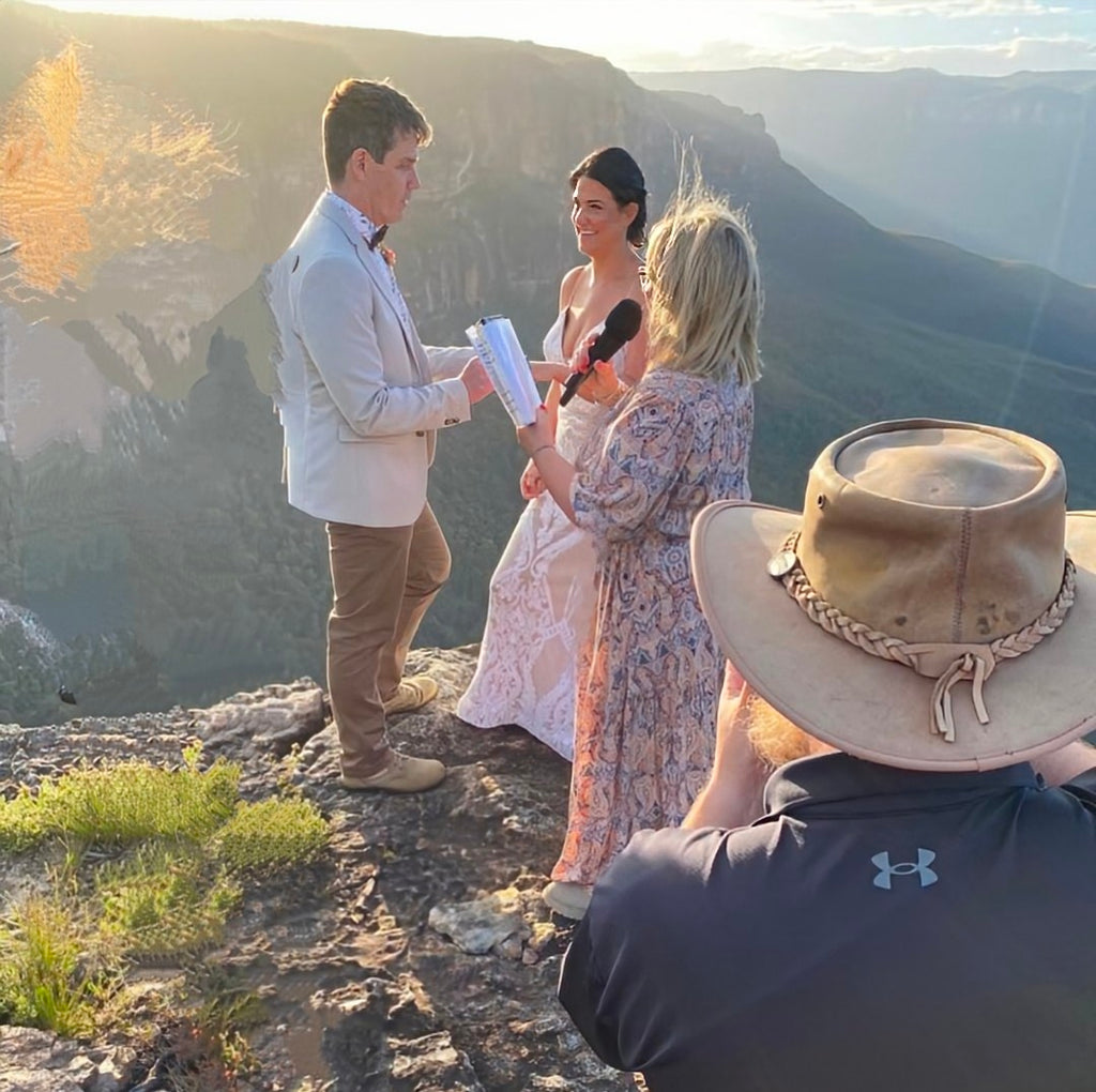Two people standing on a cliff overlooking a scenic landscape, with a third person in the foreground wearing a wide-brimmed hat.