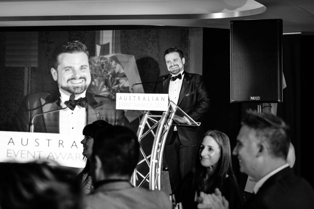 A siperimposed image of a main in formal attire standing at a podium with 'Australian Event Awards' sign, audience in the foreground.