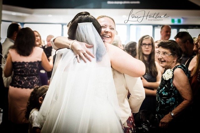 Two women embracing at a wedding reception with guests in the background.