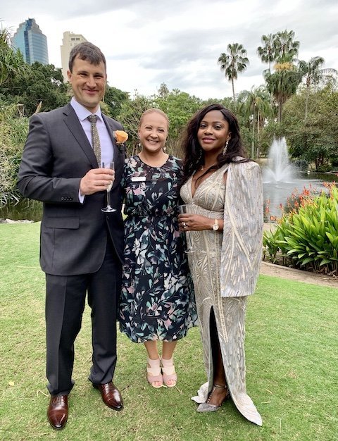 Three people posing outdoors with a fountain and trees in the background
