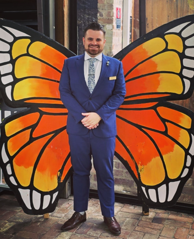 Man in a blue suit standing in front of large butterfly wings with orange, yellow, and black colors.
