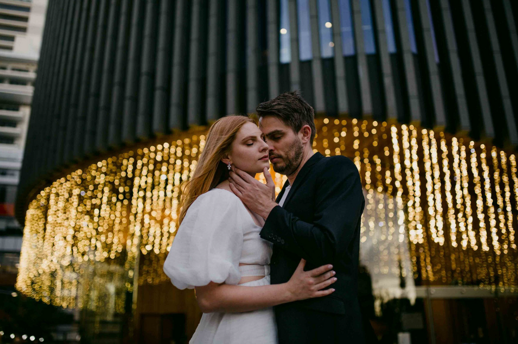 Couple embracing in front of a building with decorative lights