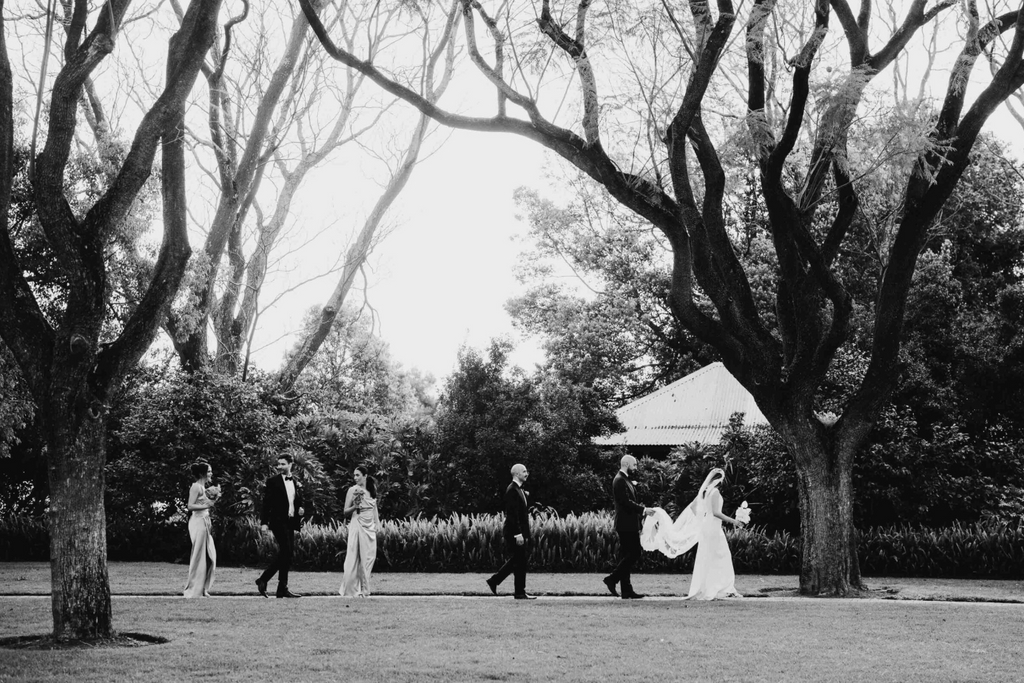 Black and white photo of a wedding party walking outdoors with trees and a gazebo in the background.