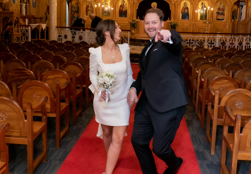 Man and woman walking down a red carpet in an elegant indoor setting with wooden chairs and decorative elements.
