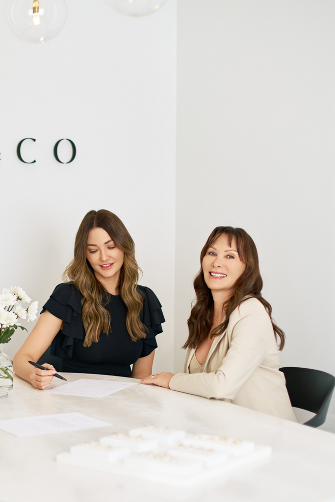 Two women sitting at a table with a white background and a partial "co" logo