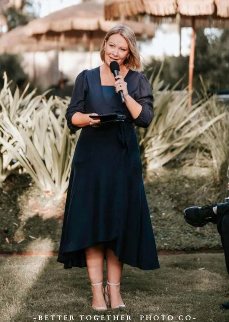Woman in a navy dress holding a microphone outdoors with plants in the background