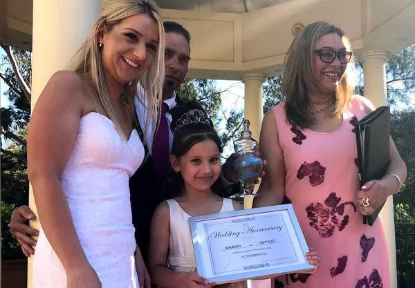 Woman in a white dress holding a 'Wedding Anniversary' certificate, standing with two other women and a child outdoors.