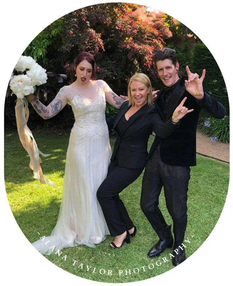 Three people posing outdoors with a wedding theme, including a bride in a white dress and two men in suits.
