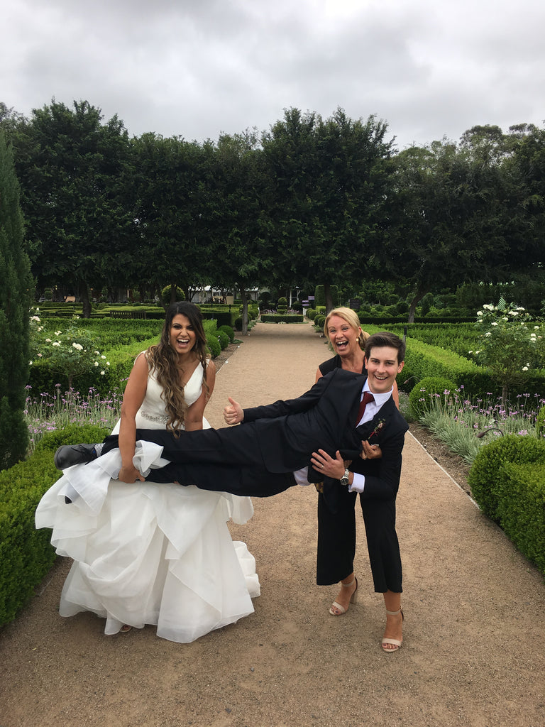 Man lifting a woman in a wedding dress with another man standing behind them, all in a garden setting.