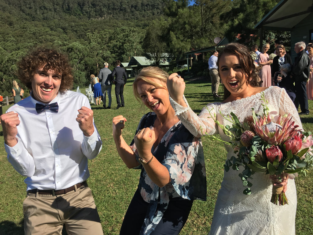 Two people celebrating outdoors with a third person holding flowers, surrounded by people and greenery.