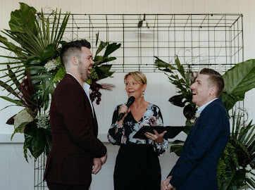 Two men standing next to a woman holding a microphone and clipboard in front of a decorative metal grid and plants.