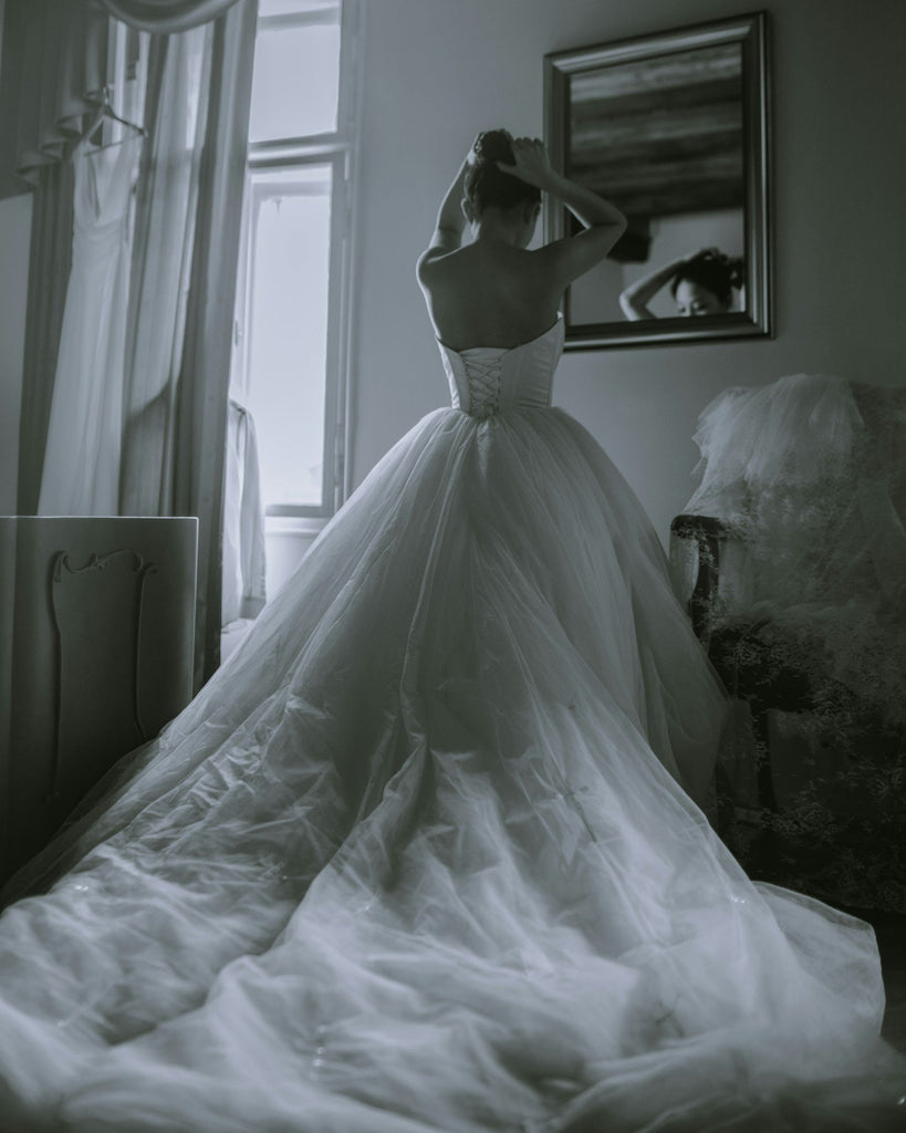 Person wearing a white wedding dress in a room with a mirror and window.
